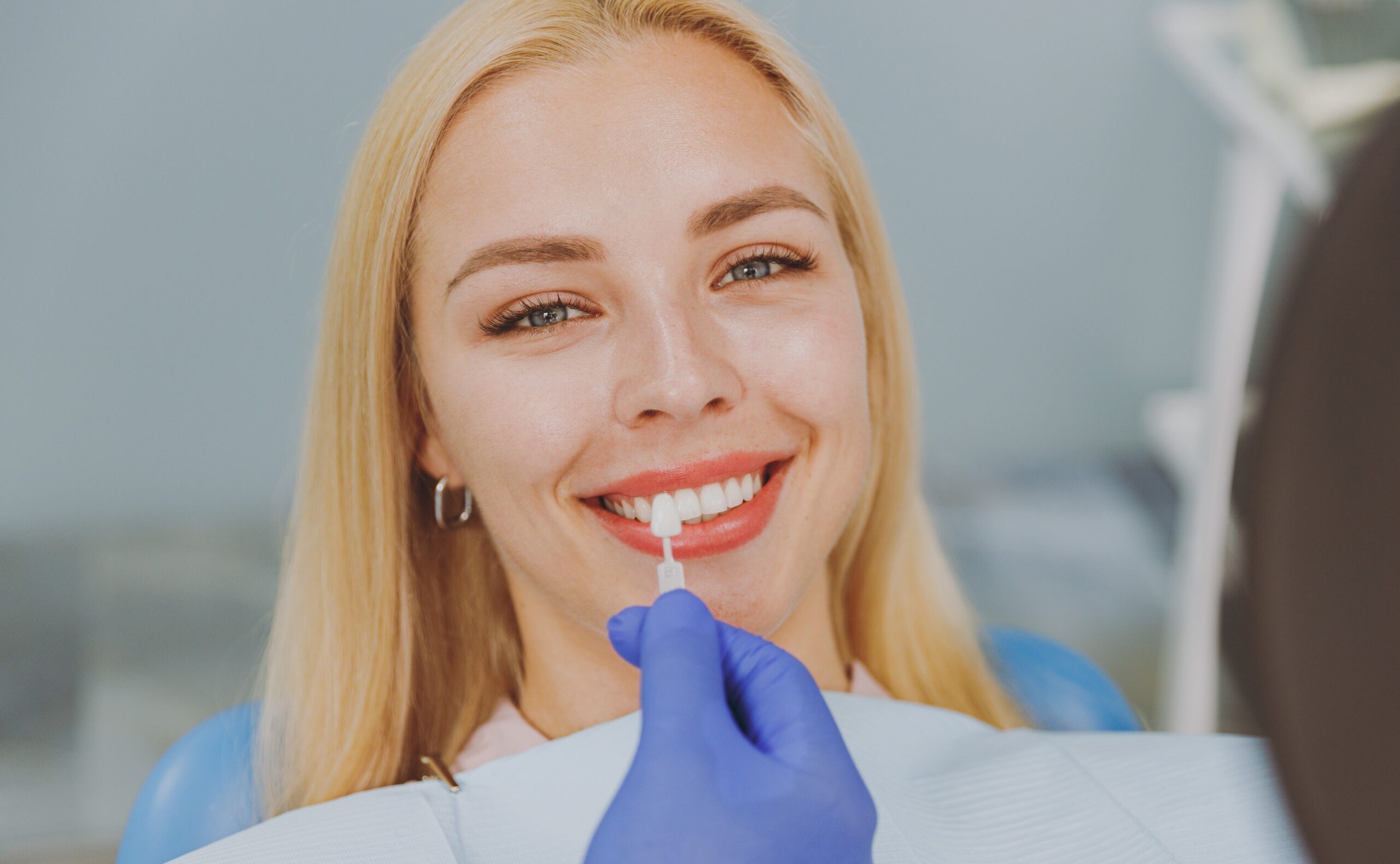 Young smiling calm happy woman choose veneers enamel color compare teeth shade with stomatologist look at result mirror sit at dentist office chair indoor modern cabinet. Enamel whitening treatment.