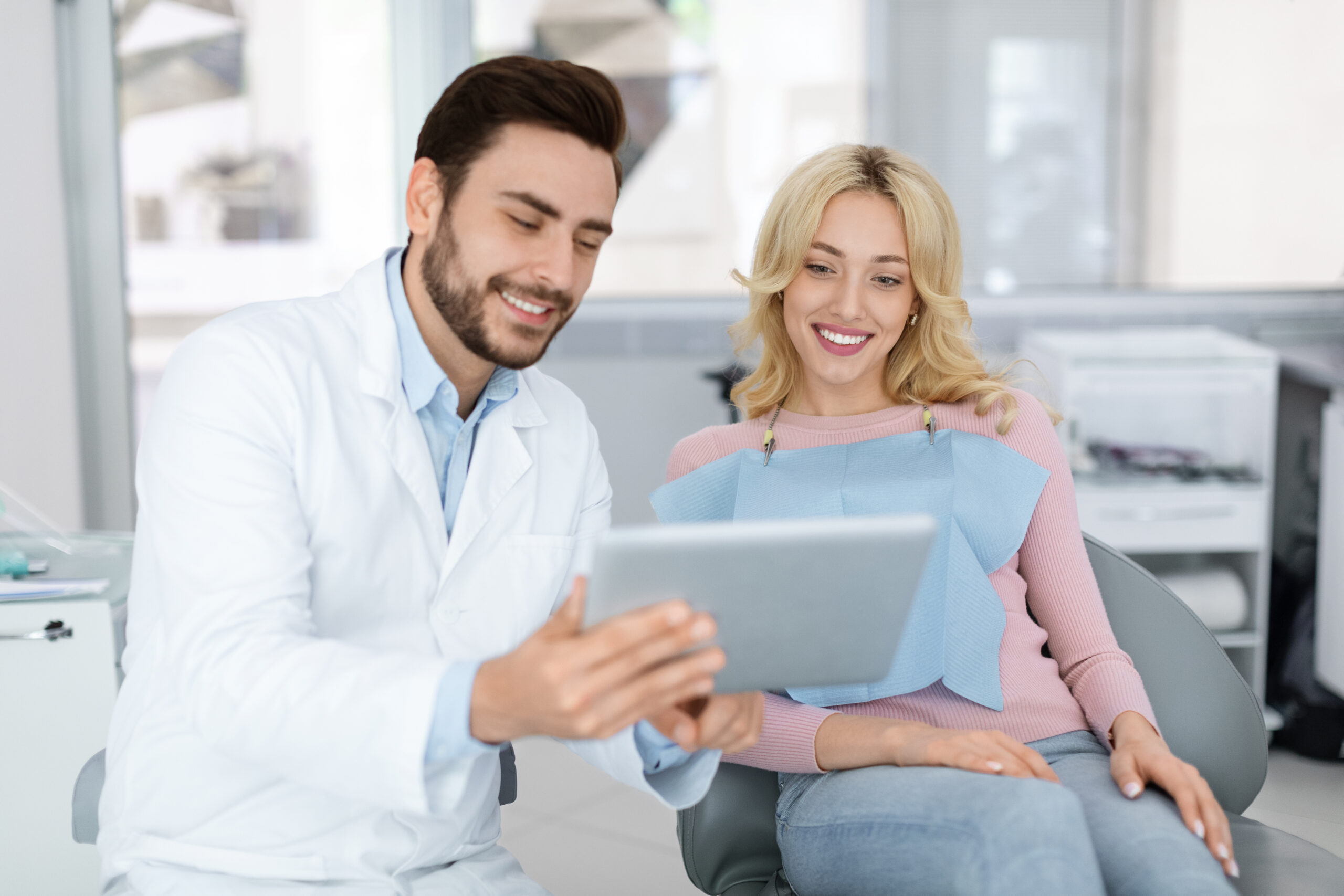 Cheerful young bearded man dentist discussing treatment method with beautiful blonde lady patient sitting at dental chair, looking at digital tablet. Modern technologies and healthcare concept