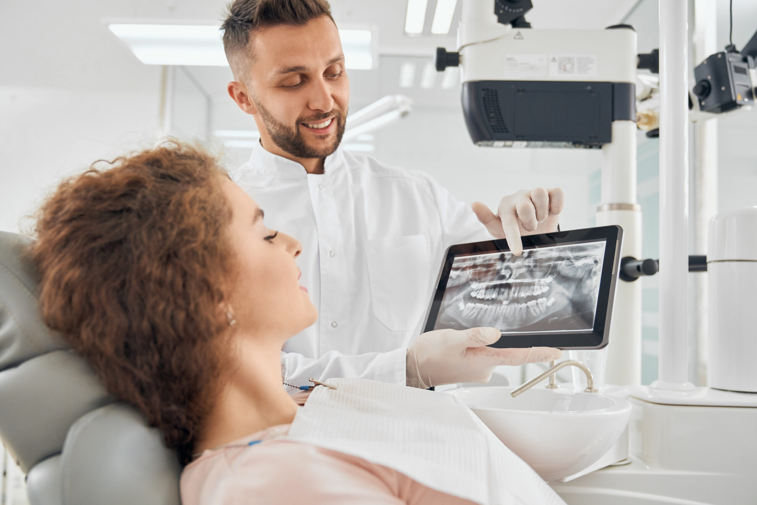Side view of beautiful girl with curly hair sitting in dental chair and looking at electronic device that holding handsome male doctor. Professional dentist showing x-ray pictures of teeth to client