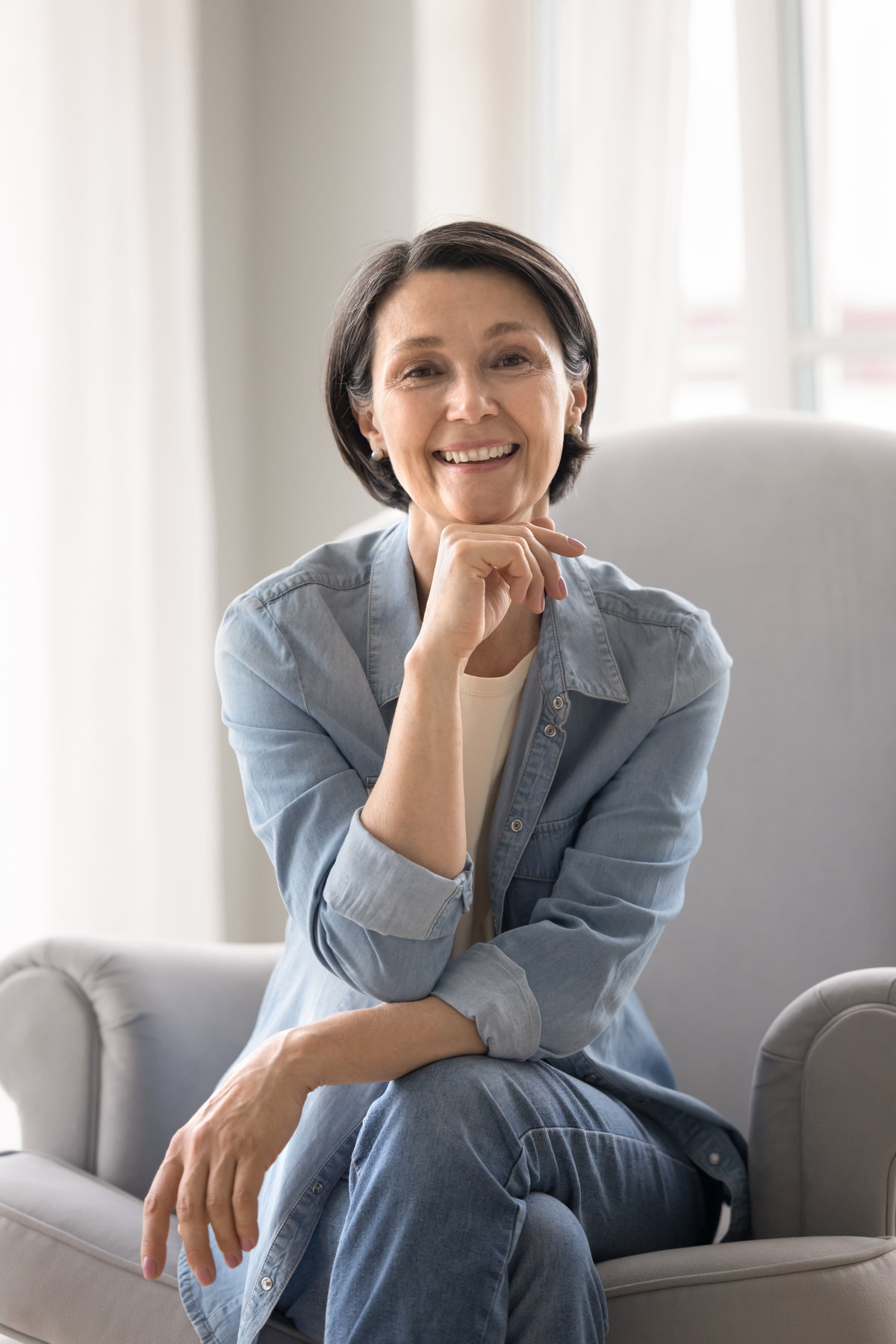 Ageing elegance. Portrait happy healthy older lady on retirement sit in cozy armchair in relaxed pose wear stylish denim clothes look at camera enjoy life with beautiful smile show optimism confidence