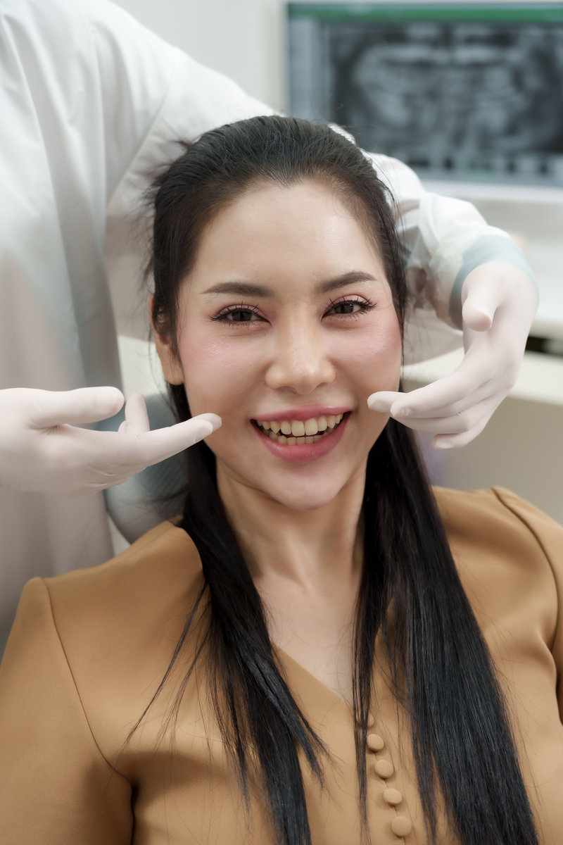 Asian adult female patient smiling after oral checkup and dental care treatment while asian adultfemaledentist wearing gloves pointing at teeth inside clean modern clinic