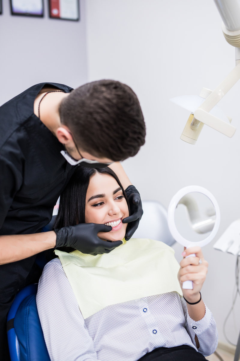 A woman is sitting in a dental office in a dental chair holding a mirror in front of her are dentists. Happy patient and dentist concept.