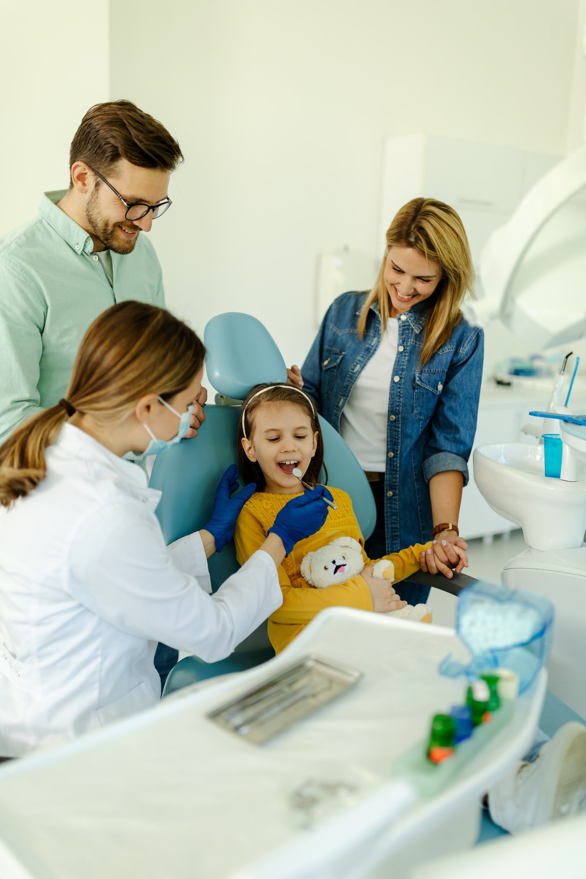 Female doctor with instruments in the hands looking girls teeth sitting in the dental chair while mother and father standing near her for support.