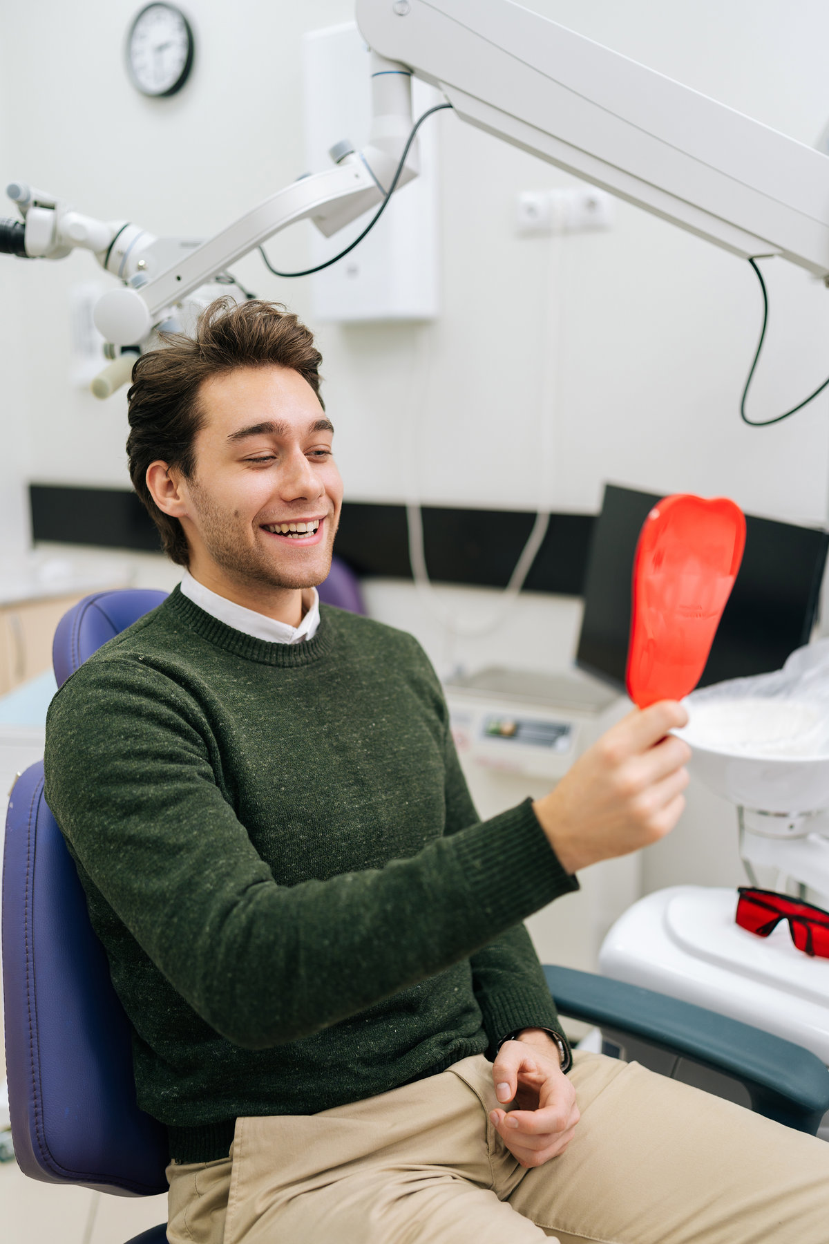 Vertical portrait of handsome cheerful young male patient smiling into mirror looking at healthy white teeth enjoying dental healthcare facilities in modern dentist office. Concept of dentistry.