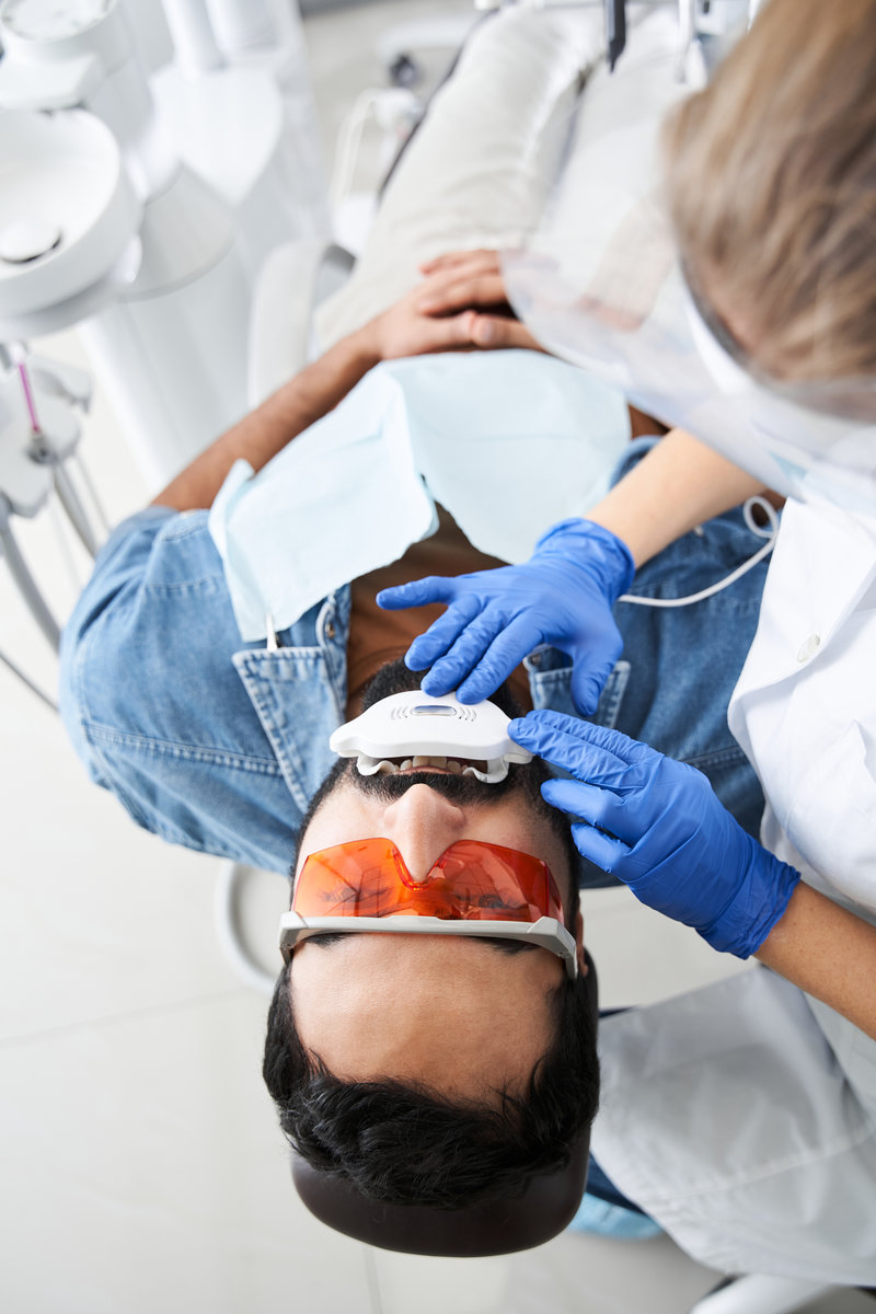 Top view of the unrecognizable dentist whitening teeth of young bearded man sitting in dental chair in modern clinic. Teeth whitening concept. Stock photo