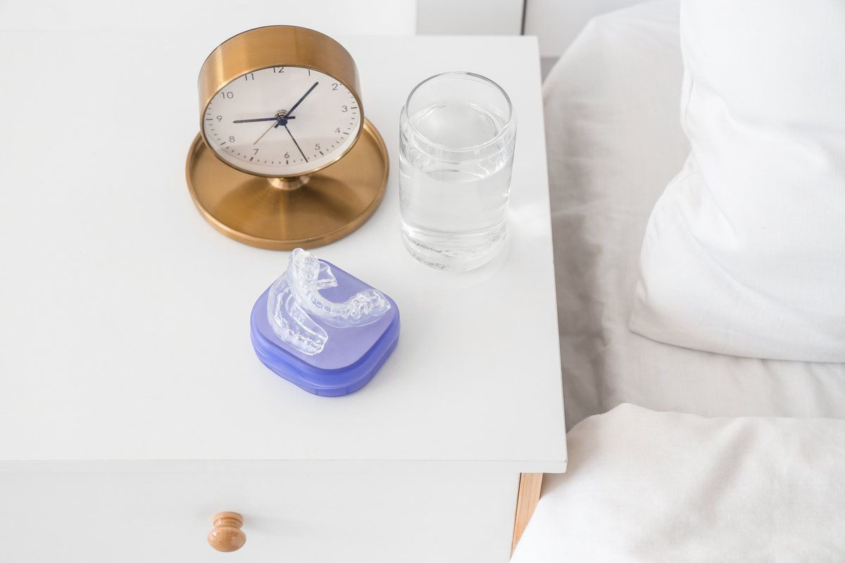 Box with occlusal splint, glass of water and alarm clock on table in bedroom, closeup